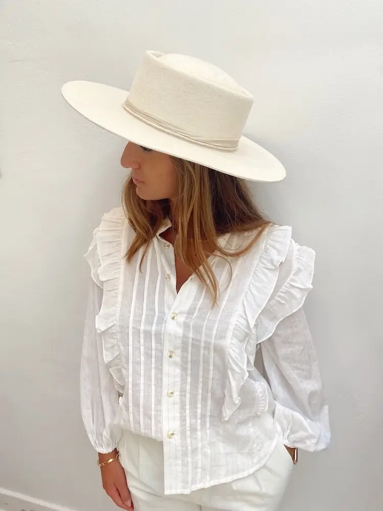 A woman, dressed in a white blouse with ruffled details and white pants, is wearing a beige wide-brimmed hat. She gazes slightly downward and to the side against a plain white background, embodying the effortless elegance of Shop Surf + Sand.
