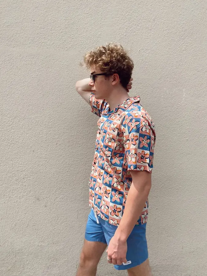 A person with curly hair is standing against a plain wall, wearing a colorful Hawaiian shirt with a floral pattern, blue shorts, and sunglasses from Shop Surf + Sand. They have one hand behind their head and are looking to the side.