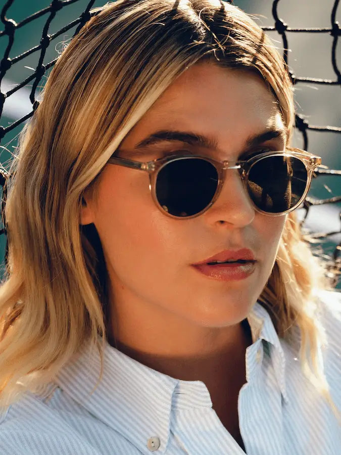 A person with long blond hair, wearing round sunglasses and a white button-up shirt from Shop Surf + Sand, is standing in front of a sunlit chain-link fence. The background is partially blurred, highlighting their face with a calm expression.
