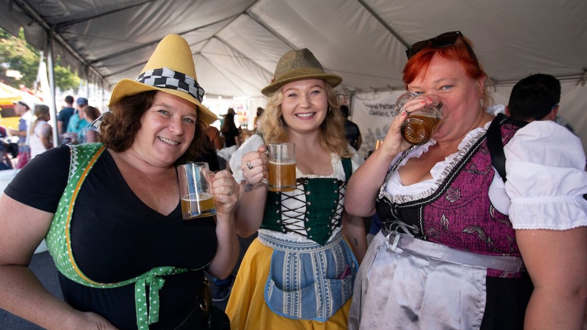 Three women dressed in traditional Bavarian attire are standing under a tent, holding and drinking beer mugs. They are smiling and appear to be enjoying themselves. The festive atmosphere makes it clear why Oktoberfest celebrations rank among the best things to do East Bay October events.