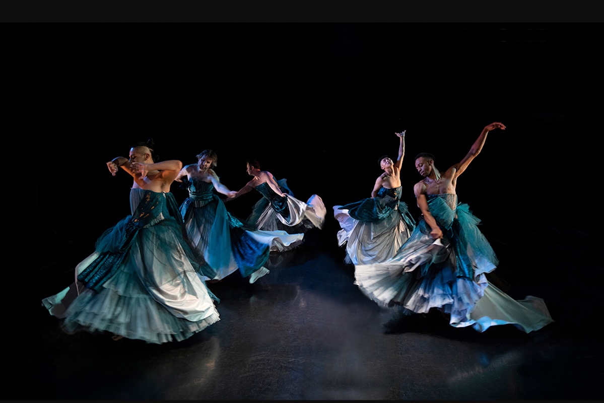 Dancers perform on a dark stage wearing flowy dresses in hues of blue and cream.