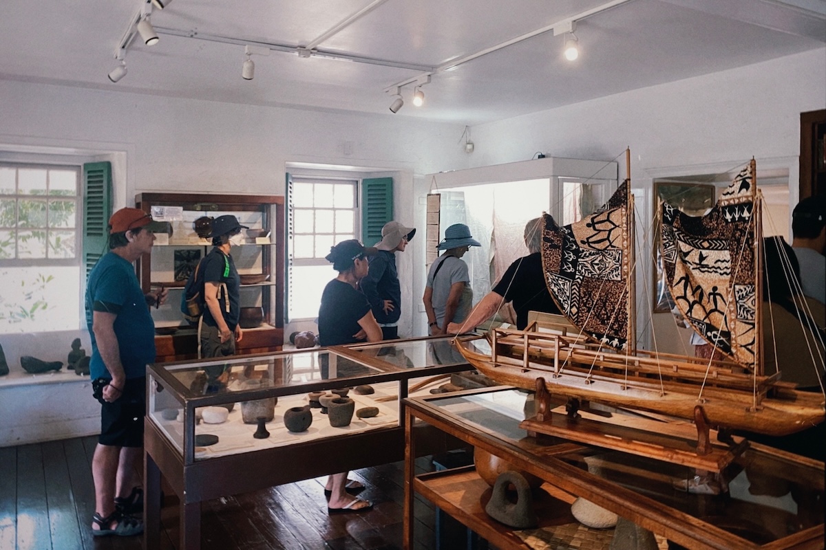A group of people walk through a museum on a tour, with dioramas of boats.