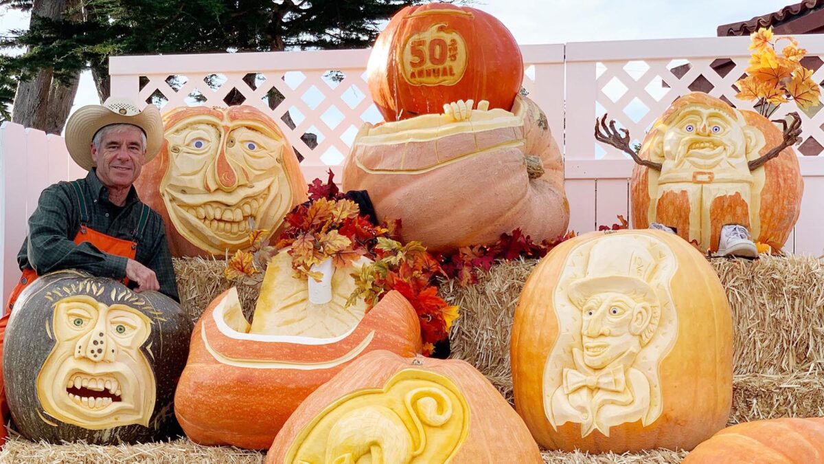 A man in a cowboy hat poses next to intricately carved pumpkins displayed on hay bales. The pumpkins feature various faces, a pirate, and a happy character. One large pumpkin at the top reads "50th Annual." Autumn leaves decorate the arrangement. One of the best things to do in South Bay this October!