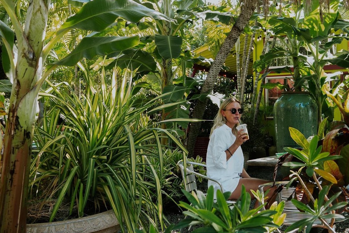 A woman wearing all white sits outside surrounded by different types of plants and greenery.