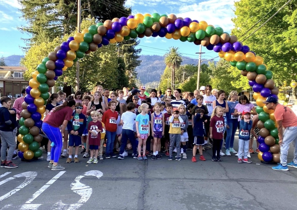 A diverse group of children and adults gather at a starting line beneath an arch made of green, gold, and purple balloons, ready for a race. Some participants wear numbered bibs. The setting is outdoors in Napa Valley with trees, a mountain, and a clear sky—one of the best things to do here this October.
