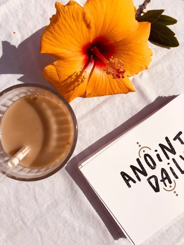 A glass of iced coffee sits on a white surface next to an orange hibiscus flower and a stack of cards. The top card reads "ANOINT DAILY" in bold, black letters, capturing the essence of Shop Surf + Sand’s relaxing vibe.