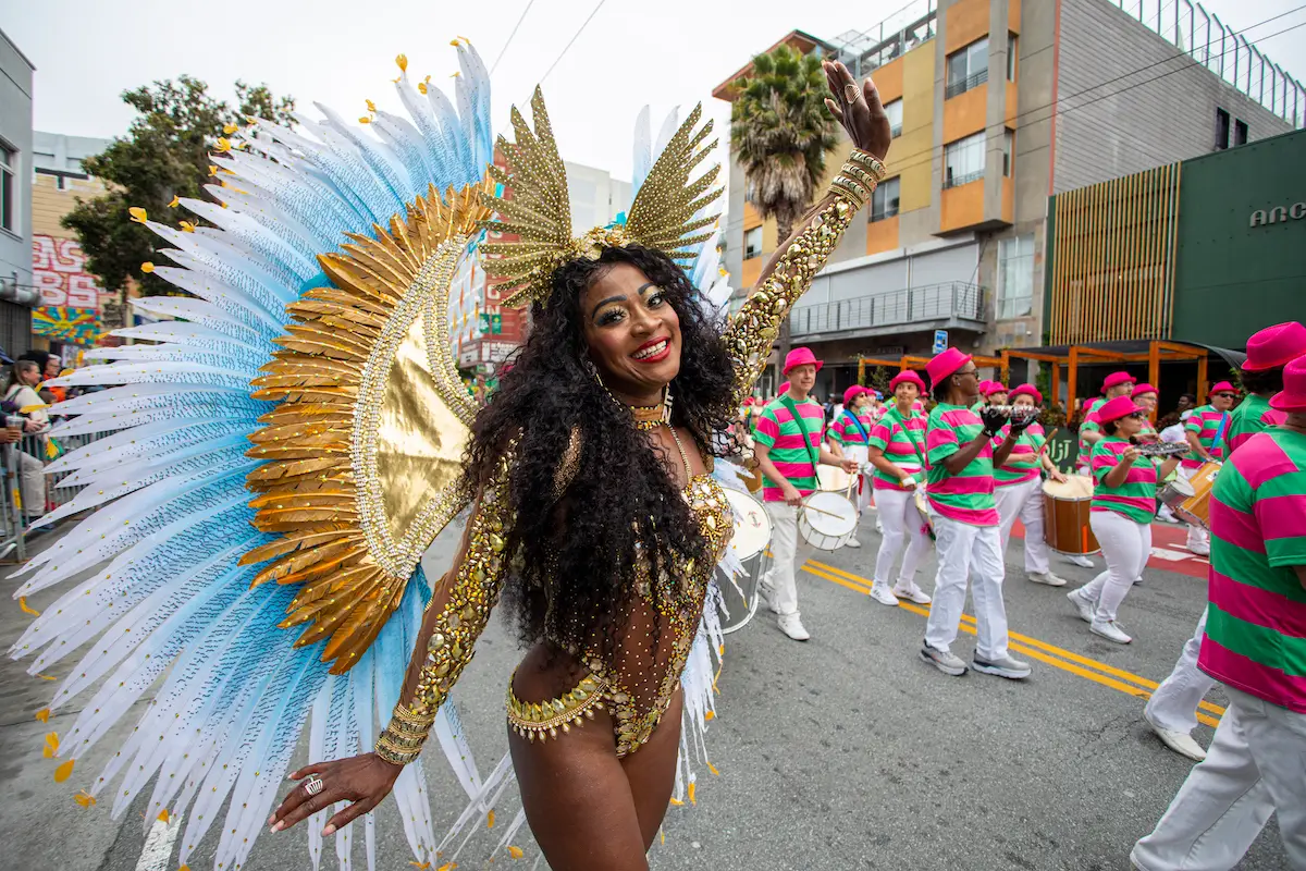 A joyful dancer in a radiant, golden feathered costume with blue accents waves during a street parade—one of the Best Things to Do in San Francisco May. She is surrounded by musicians in pink and green striped shirts and white pants, playing instruments. Urban buildings and spectators can be seen in the background.