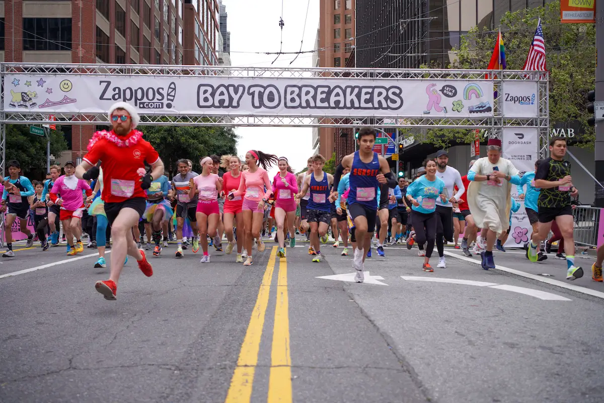 A diverse group of runners, many in colorful and whimsical costumes, dash down a city street beneath a "Bay to Breakers" banner. Tall buildings, trees, and race signage set the backdrop for one of the Best Things to Do in San Francisco in May, creating an energetic and festive atmosphere.