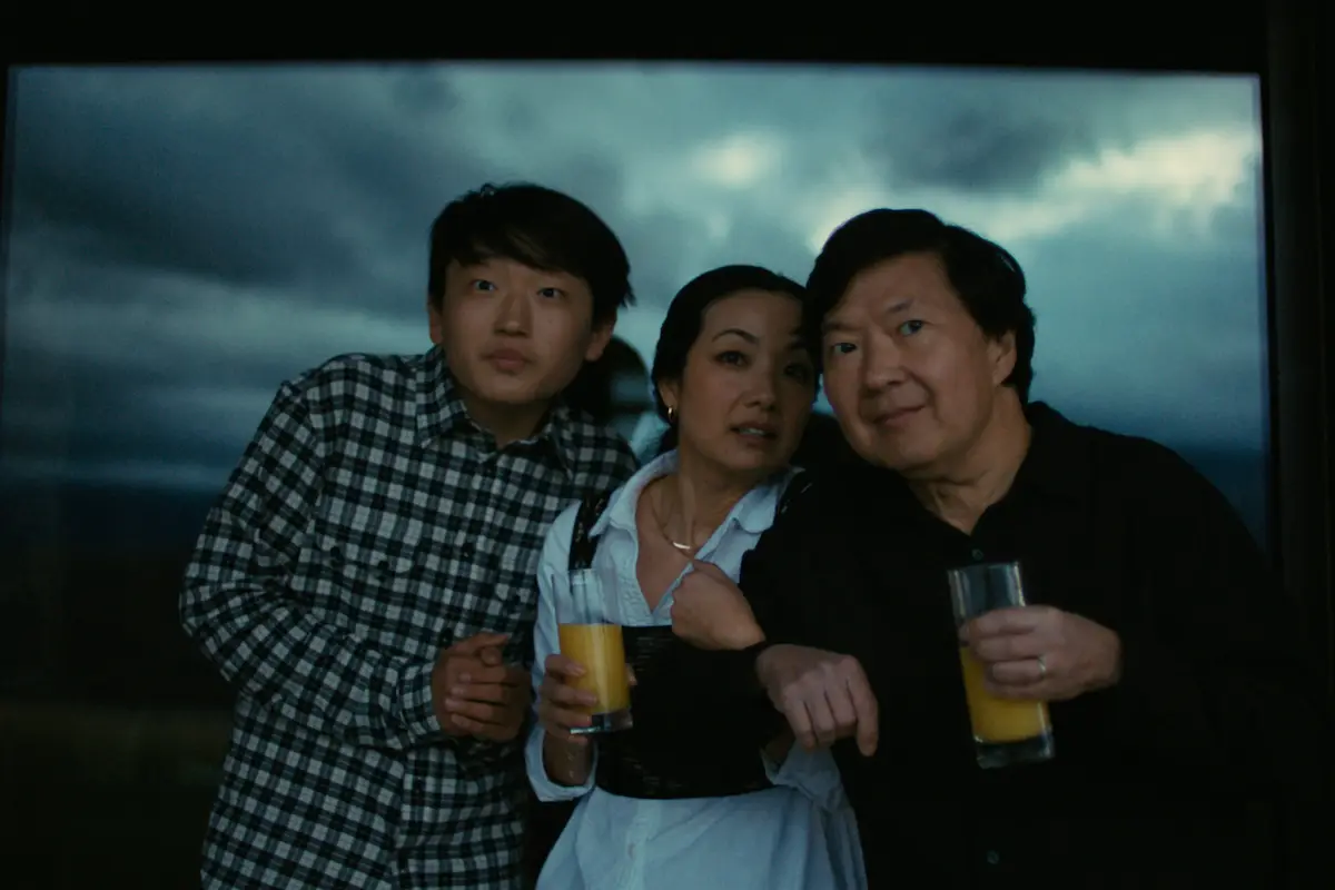 A group of three people stands together at dusk or night, holding glasses filled with a yellow beverage, likely juice. All three are looking intently at something off camera, with a dark, cloudy sky in the background. They appear to be indoors by a window, perhaps contemplating the best things to do in San Francisco this May.