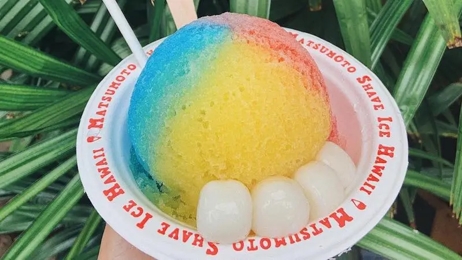A bowl of colorful shave ice in hues of blue, pink, and yellow from Matsumoto Shave Ice Hawaii, known for the best shave ice on the North Shore. The bowl is white with red text branding around the rim. Three round white mochi pieces are placed at the bottom of the bowl. A white spoon is provided.