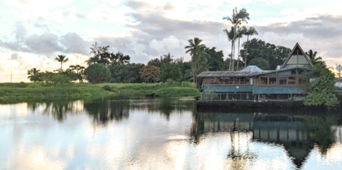 A serene tropical landscape featuring a wooden house with large windows extending over a calm body of water. Tall palm trees and lush greenery surround the house, creating the perfect view for enjoying the best dinner Hilo, Big Island has to offer, with cloudy hues of blue and grey reflected below.
