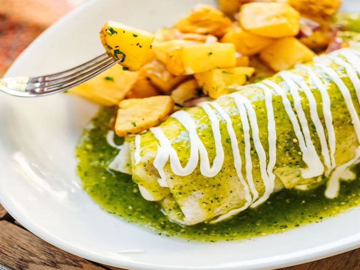 A delicious plate featuring the best breakfast in Sacramento: a burrito topped with green sauce and drizzled with white cream. On the side, there are golden, seasoned roasted potatoes, with a fork holding a piece of potato above the plate.