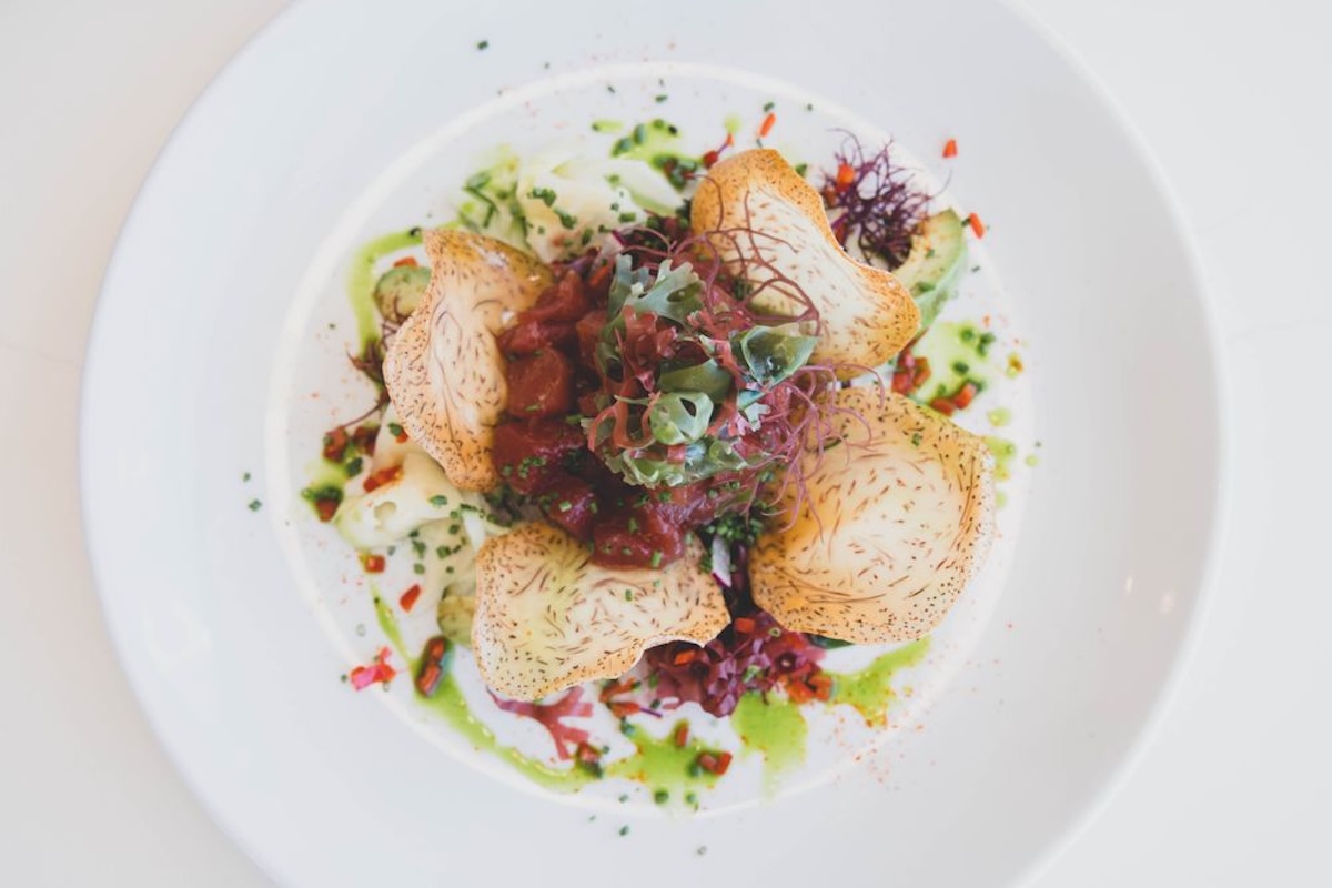 A white plate with ahi poke, taro chips, and white and green sauce for garnish.