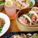 Pork bun tacos, shrimp, noodles, a cocktail and more arranged on a table.