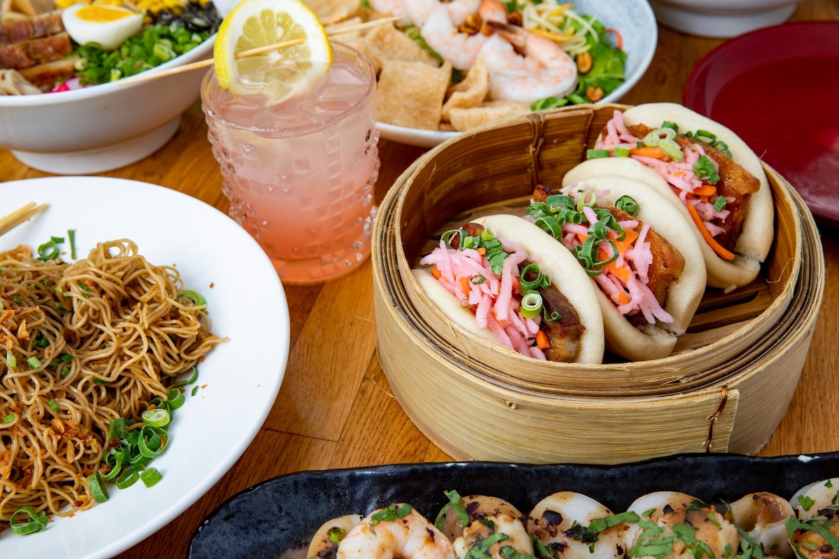 Pork bun tacos, shrimp, noodles, a cocktail and more arranged on a table.