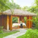 A serene outdoor massage hut with a thatched roof is set amid lush tropical greenery, exemplifying why this location is one of the best spa hotels in Kauai. The wooden walls have wide openings, offering views of the verdant surroundings as a massage therapist prepares the table with white linens.