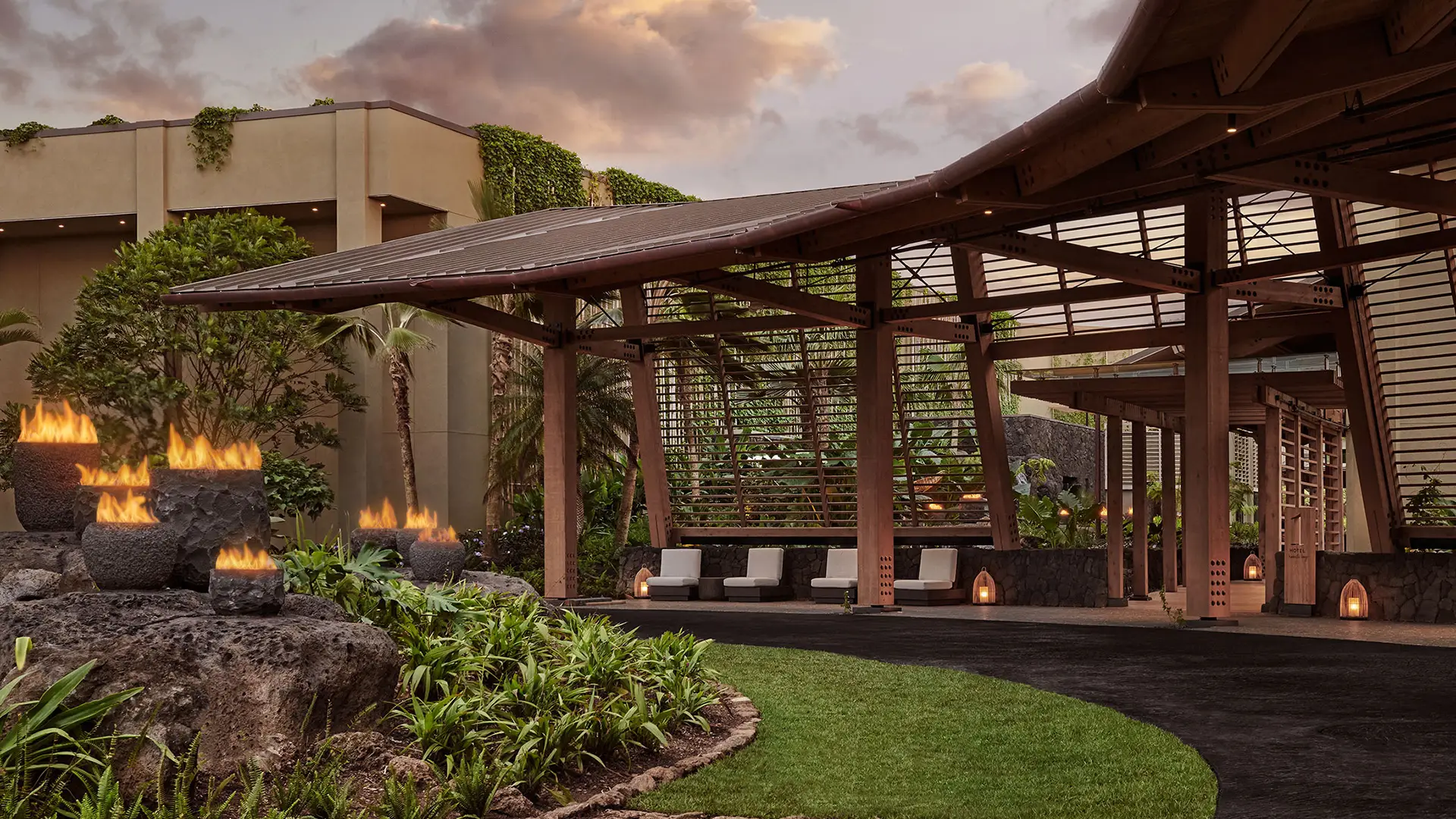 A serene outdoor seating area with a wooden pergola and modern stone fire pits, surrounded by lush greenery and tropical plants. The sky is partly cloudy, and the adjoining building is partially covered in green plants, adding to the tranquil atmosphere reminiscent of the best luxury hotels in Kauai.