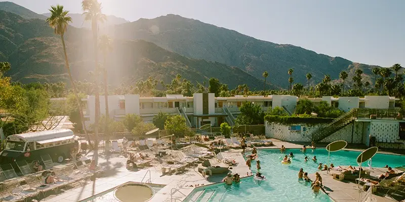 A lively outdoor pool scene with people swimming and lounging on sunbeds under a clear sky. A mountain range in the background adds to the scenic beauty. A retro van is parked nearby, and the pool area is surrounded by modern buildings and palm trees, straight out of The Ultimate Guide to Palm Springs.
