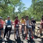 A group of eight people, all wearing helmets and casual summer clothes, are lined up on bicycles on a paved trail surrounded by trees and greenery. The sunny weather and shadows on the ground suggest it is a bright day, showcasing one of the top activities in Sonoma.