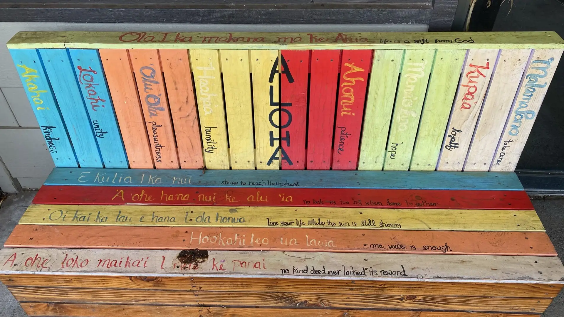 A colorful wooden bench with slats featuring Hawaiian words and their English translations. The slats read unity, humility, patience, perseverance, honesty, many hands make light work, love all you see, and kindness. The largest slat highlights the Hawaiian phrase "ALOHA.