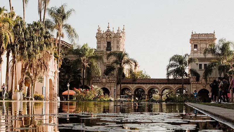 A picturesque view of a Spanish colonial-style building, evocative of Southern California Missions, surrounded by palm trees and reflected in a clear, lily-pad-covered pond in the foreground. People walk and gather near the building, enjoying the sunny day.