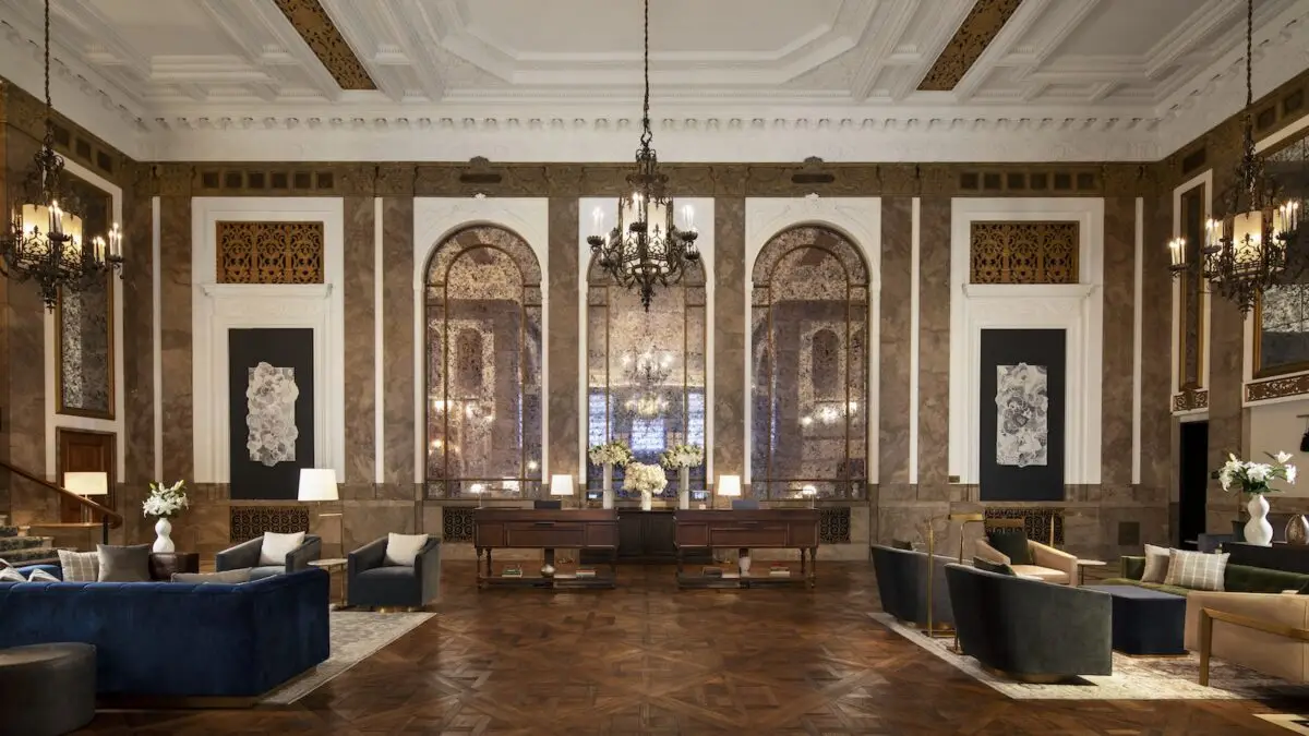 The Beacon Grand hotel lobby boasts high ceilings, ornate chandeliers, and tall arched windows. Its elegant furniture includes couches, chairs, and side tables with lamps. Decor features large mirrors, intricate wall designs, and tasteful floral arrangements.