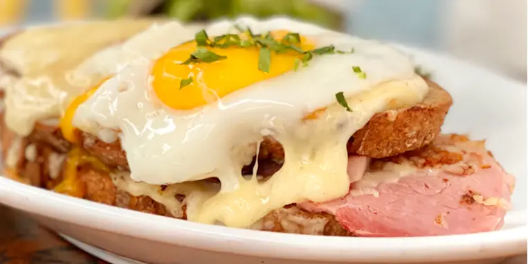 A close-up of a plate featuring a croque madame sandwich. It consists of slices of ham, melted cheese, and a thick layer of béchamel sauce, topped with a sunny-side-up egg sprinkled with fresh herbs. The dish is placed on a white plate – truly the best breakfast Sonoma County has to offer.
