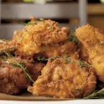 A plate of crispy fried chicken pieces garnished with fresh herbs, set on a parchment-lined surface. The golden-brown coating appears crunchy, reminiscent of the best dinner in Napa Valley. In the background, a blurred wooden chair and piece of furniture complete the cozy scene.