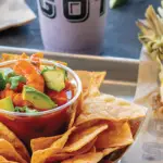 A tray with a bowl of nachos topped with diced avocado, salsa, and lime wedges. Beside it, a piece of grilled corn on the cob is coated with spices and a white crumbly topping. A drink in a cup is in the background, making for the best lunch experience you could have in Napa Valley.