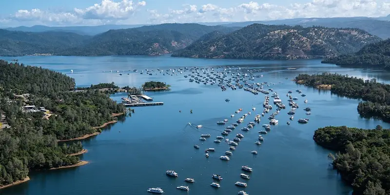 A scenic aerial view of Lake Oroville, a perfect summer getaway surrounded by lush green hills and mountains. Numerous boats are anchored in the water, forming lines that extend towards the shore. A marina is visible to the left, with docks and buildings along the shoreline.