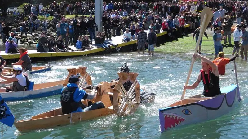 Participants race in creatively designed boats during a fun, colorful event on water, with a large, cheering crowd on the shoreline. One boat is shark-themed, and another appears to have a wooden structure. People are paddling energetically to move their boats. It's among the best things to do in Wine Country this February!
