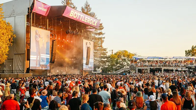 Image of a large crowd at an outdoor music festival in front of a stage with a "Bottlerock" banner. Two large screens on either side of the stage show a performer singing. Trees, lighting equipment, and a structure with seated viewers are visible in the background—one of the best things to do in Wine Country January.