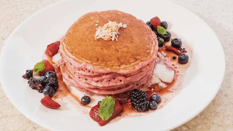 A stack of pink pancakes is topped with shredded coconut and surrounded by fresh berries, including strawberries, blueberries, blackberries, and a garnish of mint leaves, creating a vibrant breakfast reminiscent of Oahu's tropical mornings on a white plate with a light-colored background.