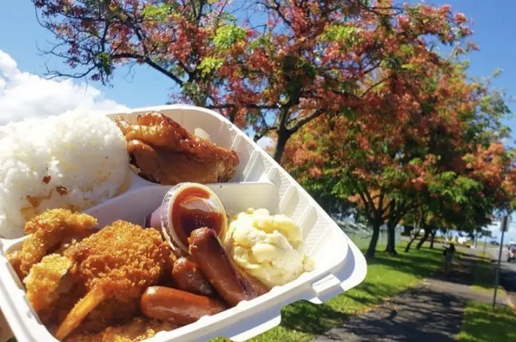 A takeout container holding rice, fried chicken, sausage, macaroni salad, and a piece of grilled meat is being held up in the foreground. The background shows a sunny park with trees featuring autumn foliage and a clear blue sky—enjoying the best Hawaiian food on the Big Island.