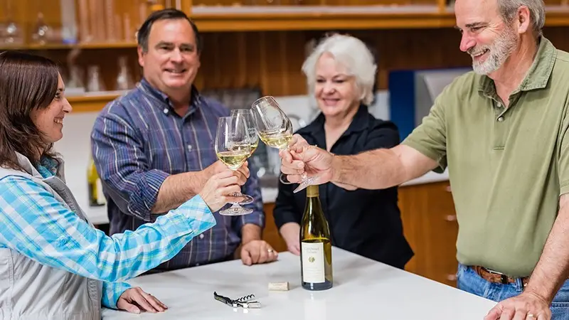 Four people clink glasses of white wine together in a cheerful toast, celebrating one of the best things to do in Wine Country. They are standing around a kitchen island, with a bottle of white wine and a corkscrew on the counter. Smiles indicate they’re enjoying the moment.