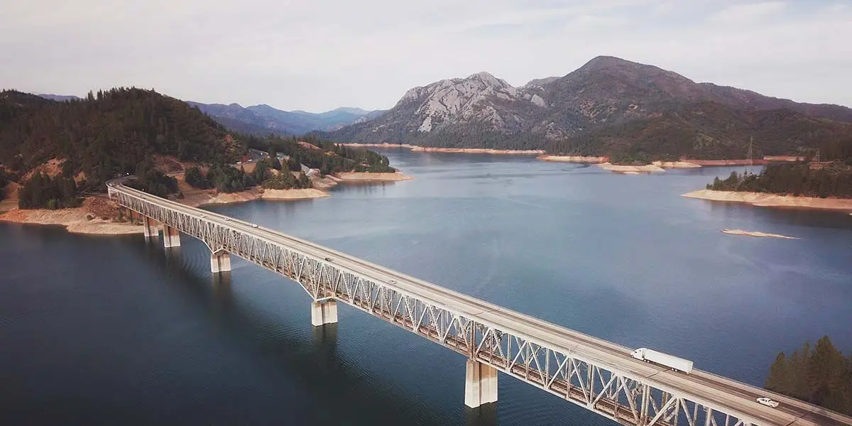 An aerial view of a long bridge spanning a wide body of water, surrounded by hilly terrain and dense forests in what could be part of a California road trip near Lake Shasta. Mountains rise in the background under a lightly overcast sky, and a white truck is seen crossing the bridge.