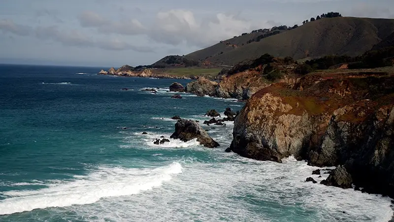 A coastal landscape featuring rugged cliffs and rocky shores with waves crashing against them, reminiscent of Monterey. The ocean is deep blue, and the sky is partially cloudy. Rolling hills with sparse vegetation can be seen in the background, offering a plethora of things to do and explore.