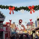 A bustling street decorated with festive garlands featuring red bows and white snowflakes captures the essence of the holiday spirit. The lively scene, filled with people enjoying the bright atmosphere, epitomizes one of the best things to do on the Monterey Peninsula in December. Various shops line both sides, adding to the vibrant ambiance.
