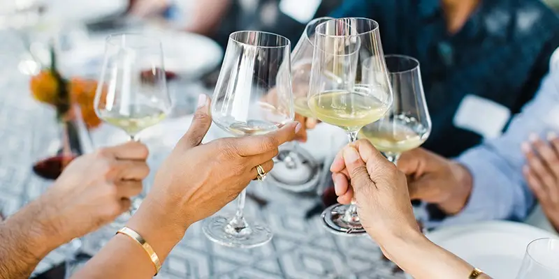 A group of people toasting with white wine glasses around a table with a patterned tablecloth. Some hands wear rings and bracelets. The background is blurred, focusing on the hands and the glasses—a perfect snapshot of the Best Things to Do in Napa Valley This October.
