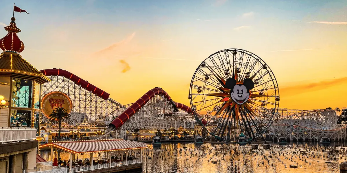 A vibrant amusement park at sunset features a large Ferris wheel with a Mickey Mouse face, colorful roller coasters, and various lit-up attractions. The sky is orange, and the reflections of the rides and lights shimmer on the water below, creating an enchanting scene straight out of the Ultimate Guide to Disneyland.