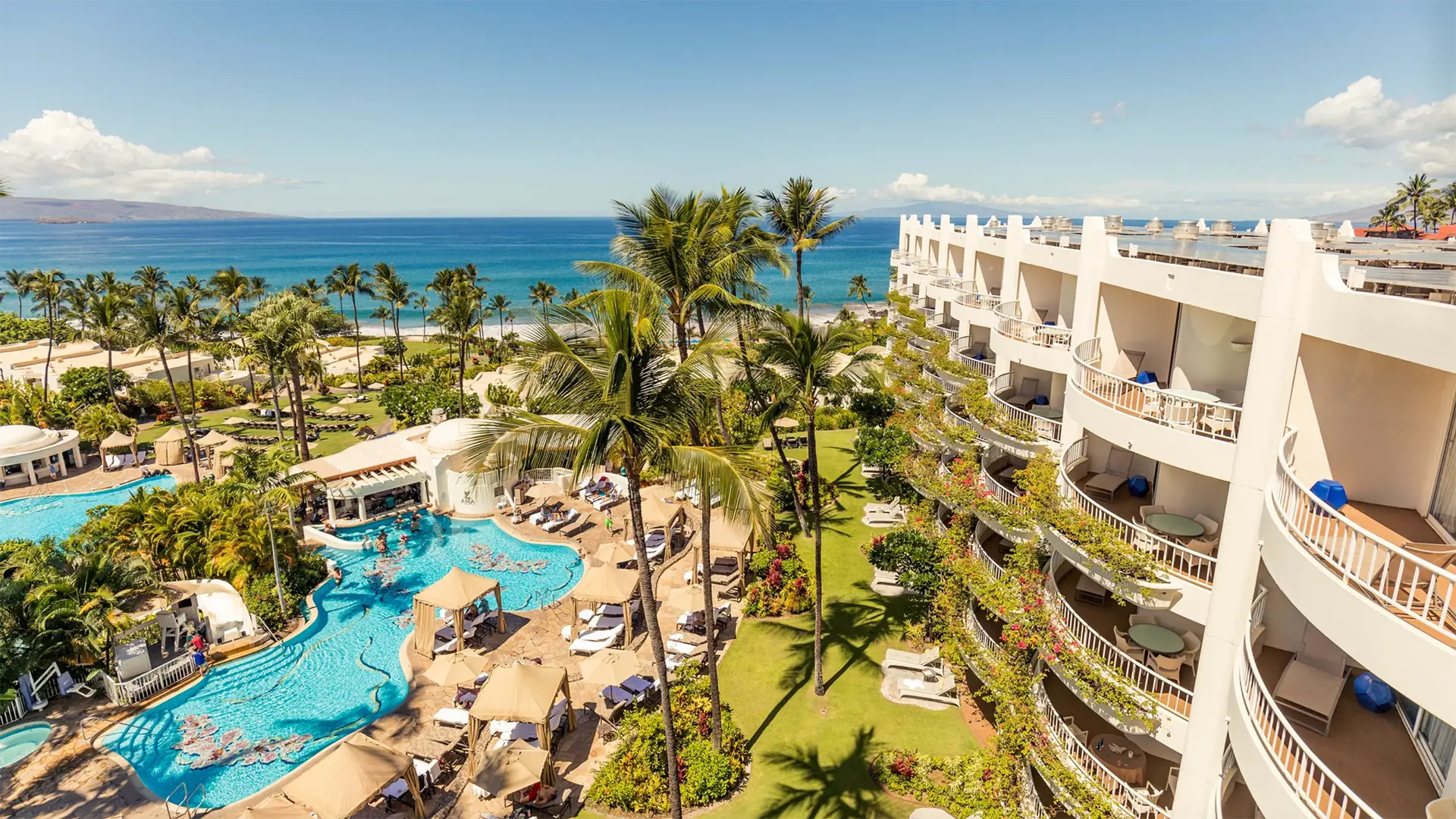 Aerial view of a luxurious beach resort with white buildings, lush greenery, and numerous palm trees. The resort features an extensive pool area with lounging chairs and umbrellas. Beyond the resort, there's a beautiful sandy beach and a blue ocean under a clear sky—one of the best group hotels Maui offers.
