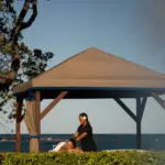 A person is receiving a massage under a beige gazebo by the ocean at a luxurious hotel spa. There are trees on the left side, and the sky is clear with a light haze. The massage therapist is standing, and greenery surrounds the gazebo. This serene escape is one of Big Island's finest offerings.