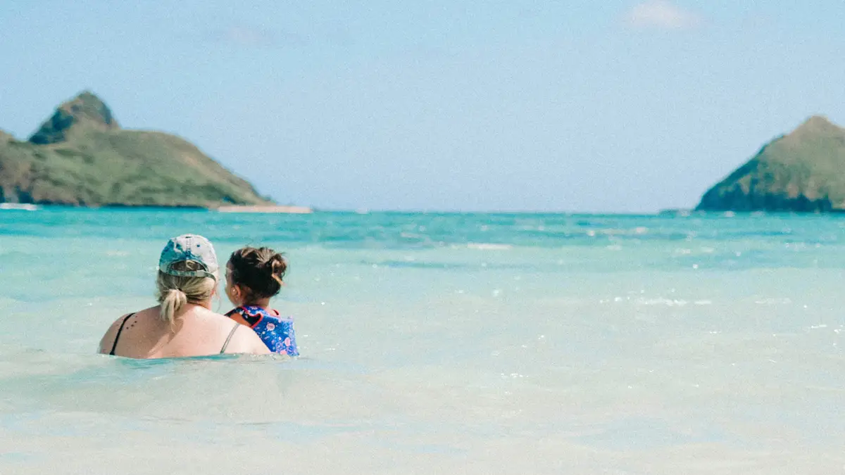 A woman and a child enjoy the clear, shallow water at a family-friendly beach on Oahu. Both are seen from behind, with the woman wearing a baseball cap and the child in a swimsuit. In the distance, two small islands or rocky hills are visible under a clear blue sky.