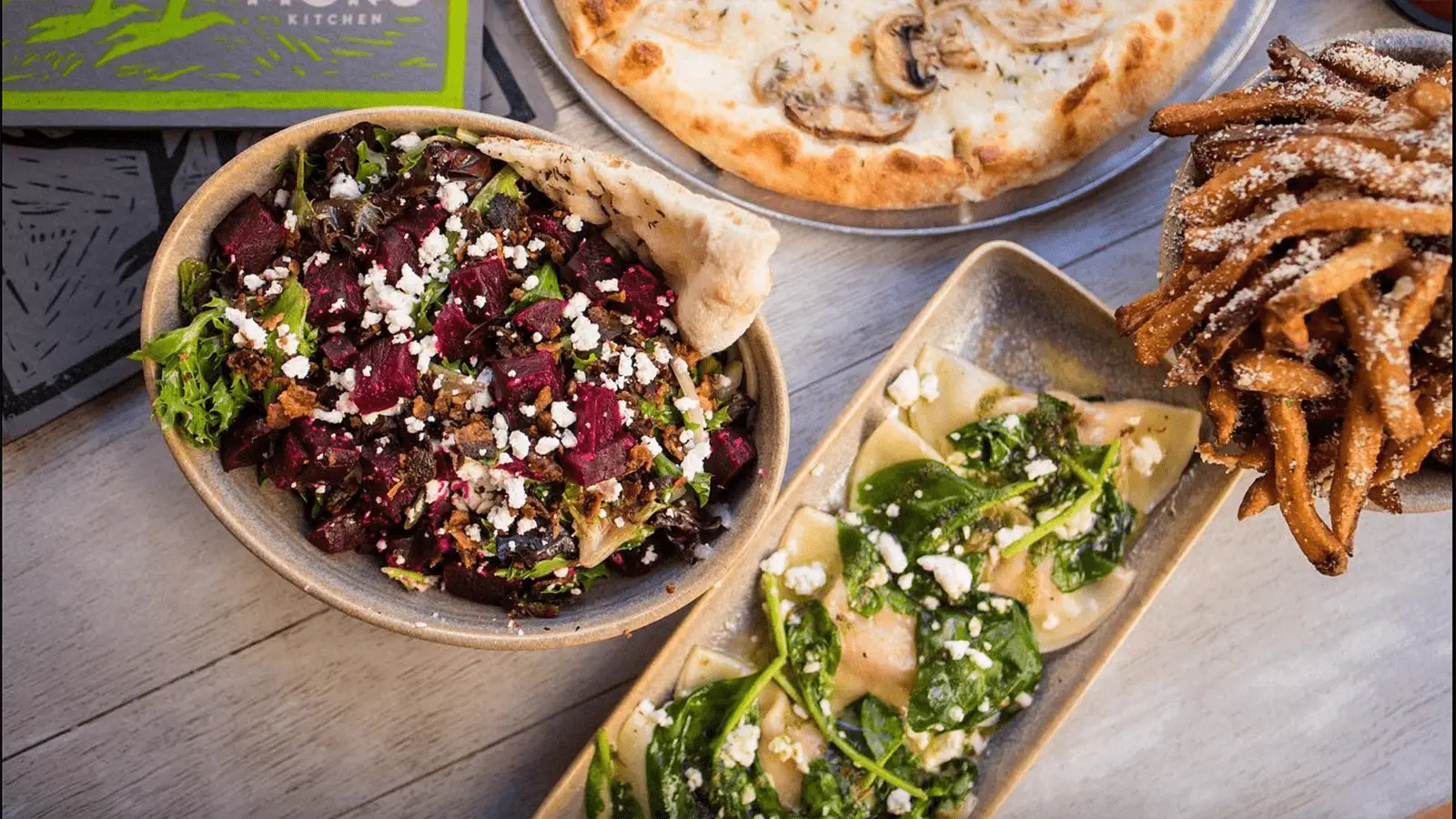 A variety of dishes on a table, perfect for the best lunch in Waikiki, Honolulu, including a bowl of salad with beets and feta cheese, a rectangular plate with spinach and cheese ravioli, a plate of seasoned fries, and a pizza topped with mushrooms.