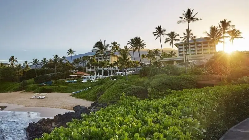 A tropical beach resort at sunrise, featuring several buildings surrounded by lush greenery and tall palm trees. This prime example of one of the best family hotels in Maui offers a sandy beach with lounge chairs near the shoreline. The sun rises behind the resort, casting a warm glow.