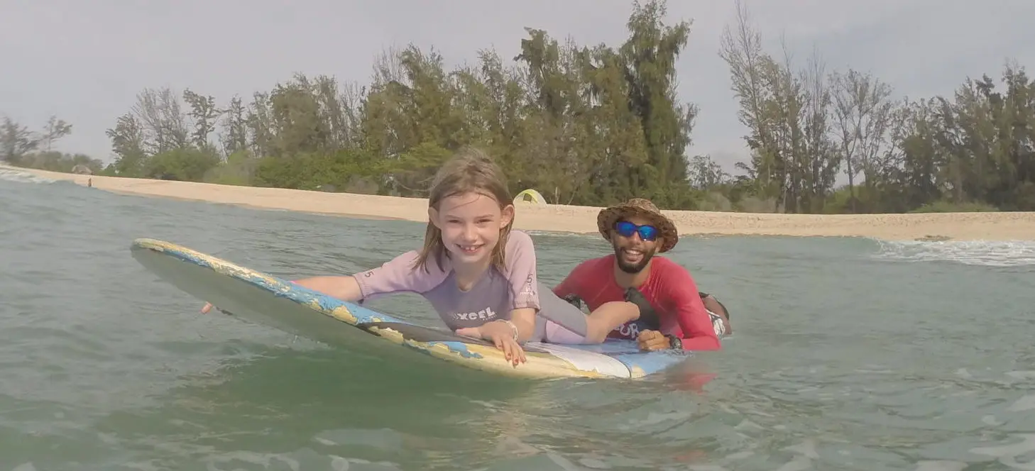 A young girl, smiling broadly, lies on a surfboard in the water with the assistance of an adult man wearing a hat and sunglasses. They are near the shore with trees and a sandy beach in the background—truly one of the best surf lessons on the west side of Oahu.