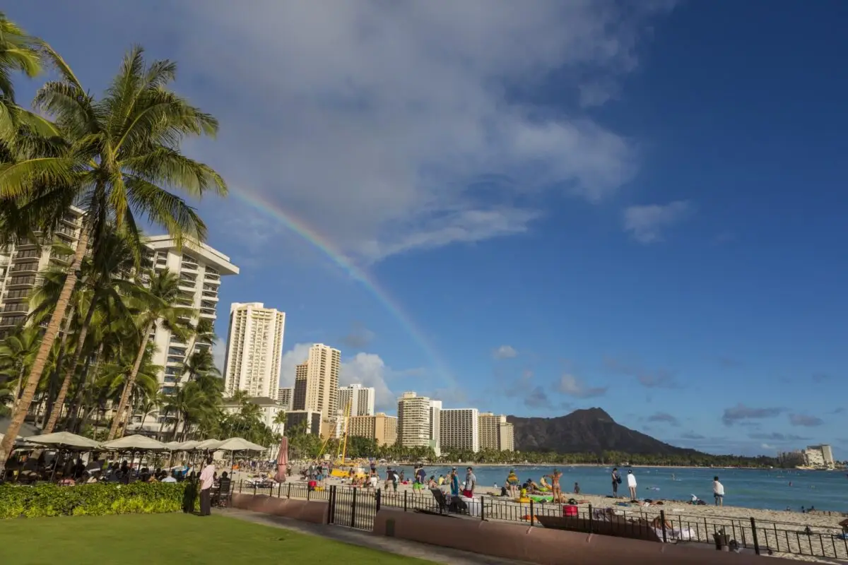 A vibrant Waikiki Beach scene with several people strolling and lounging, surrounded by tall palm trees and high-rise buildings. There is a rainbow arc in the sky above and a distant mountain in the background, all under a bright blue sky with scattered clouds.