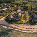 Aerial view of a beachside village, with rows of houses surrounded by greenery. The sandy beach features gentle waves and a mix of grass and rock sections. The landscape includes dense foliage and additional houses in the background, showcasing some of the best oceanfront hotels Kauai has to offer.