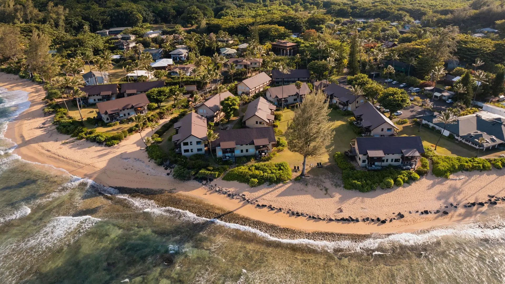 Aerial view of a beachside village, with rows of houses surrounded by greenery. The sandy beach features gentle waves and a mix of grass and rock sections. The landscape includes dense foliage and additional houses in the background, showcasing some of the best oceanfront hotels Kauai has to offer.