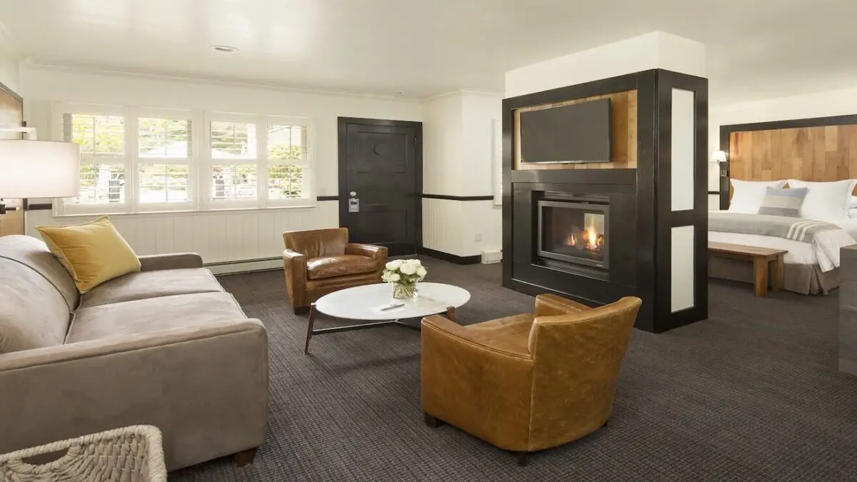 A modern living room with a beige sofa, two brown leather chairs, and a white round coffee table adorned with a vase of flowers. A fireplace with a mounted TV separates the living area from a bedroom featuring white linens. This space rivals the best romantic hotels on the Monterey Peninsula.